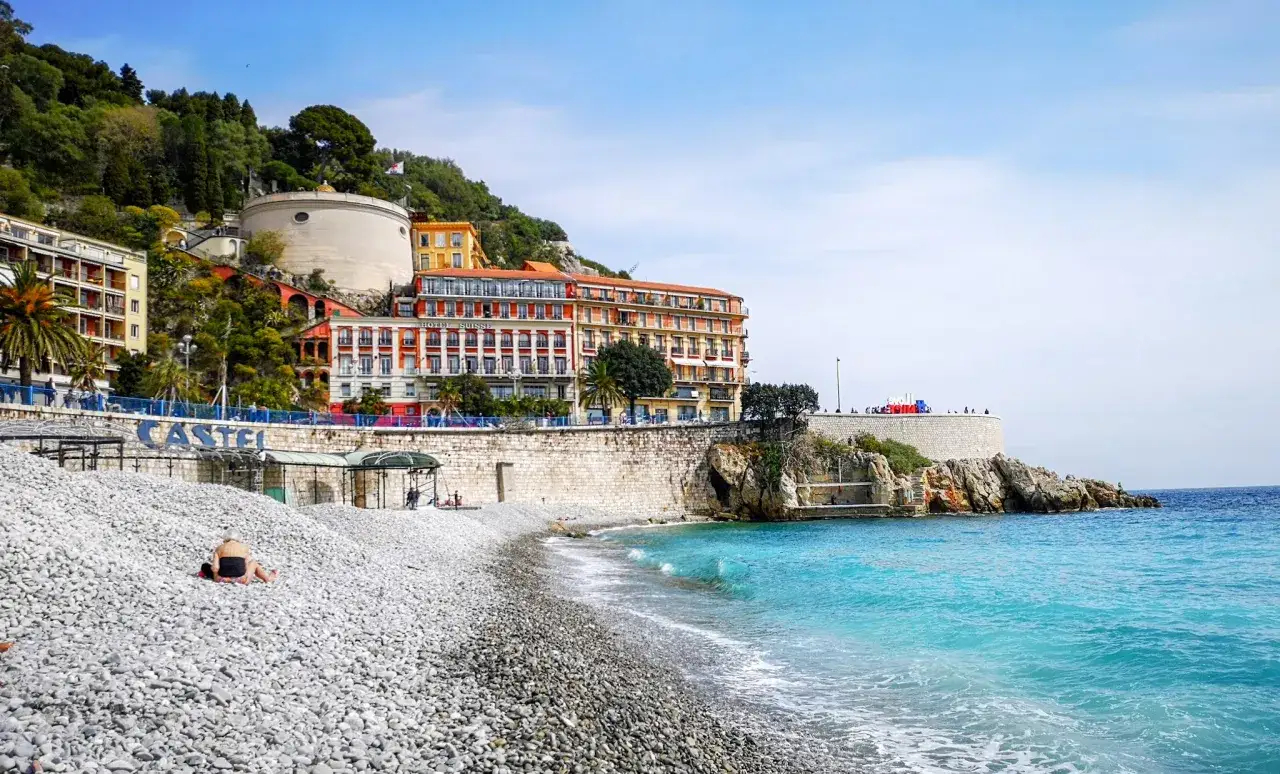 Kamienista plaża w Nicei, z widokiem na turkusowe morze i kolorowe budynki na wzgórzu. Idealne miejsce, by poczuć klimat Lazurowego Wybrzeża. Nicea co zobaczyć? To właśnie!
